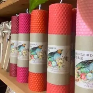 A row of tall, hand-rolled beeswax candles in shades of red, orange, and pink neatly displayed on a wooden shelf. Each candle features a kraft paper label with colorful floral artwork and a bird illustration, representing the brand 
