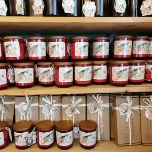 Shelves filled with red glass jar candles and kraft gift boxes from “The Songbird and The Bee,” all labeled with floral bird illustrations and decorative tags.