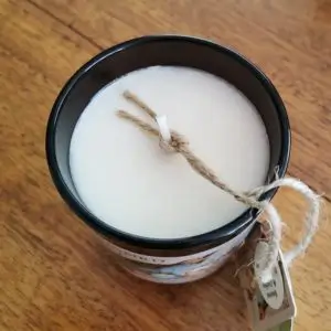 Top view of a white candle in a black glass jar with a jute string tied around the wick, featuring a “The Songbird and The Bee” label and a hanging tag on a wooden surface.