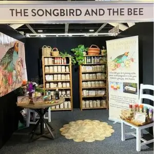 Trade show booth for “The Songbird and The Bee” featuring candle displays, promotional banners, and a round wooden table with products, set against a black backdrop with floral decor.
