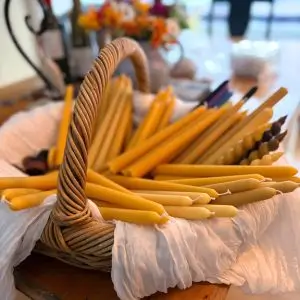 A rustic wicker basket lined with white cloth filled with an assortment of hand-dipped beeswax taper candles in various shades of golden yellow. The candles vary slightly in size and texture, and a soft-focus background shows a cozy, artisan setting with flowers and wine bottles.