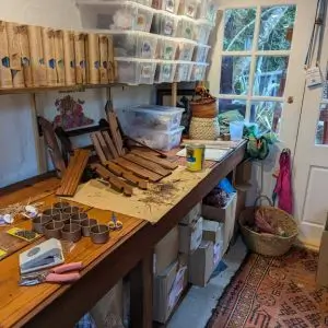 A cozy, rustic workspace with a long wooden workbench filled with craft supplies, including rolled beeswax candle packaging, cylindrical candle holders, scissors, labels, and wood stain. Wooden display stands are being assembled. Shelves above the bench are stocked with clear plastic bins containing colorful floral-themed products. A window and door let in natural light, and the space includes baskets, boxes, and a patterned rug on the floor.