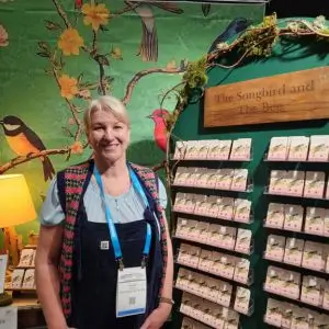A smiling woman stands in front of a colorful booth for “The Songbird and The Bee.” The display wall is decorated with a lush green floral mural featuring birds and branches. A wooden sign with the brand name hangs above rows of earring cards, each featuring the brand’s signature bird and floral artwork. The woman wears a name badge and a cheerful expression, suggesting a tradeshow or exhibition setting.