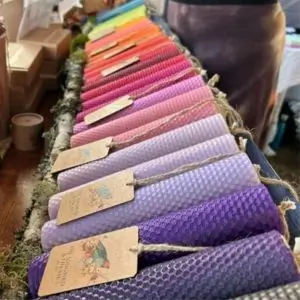 A colorful display of rolled beeswax candles arranged in a gradient from purples to pinks, reds, and oranges at a market stall. Each candle is wrapped with a branded tag from “The Songbird and The Bee.” The candles are laid out neatly on a moss-lined tray, with other products and a vendor partially visible in the background.