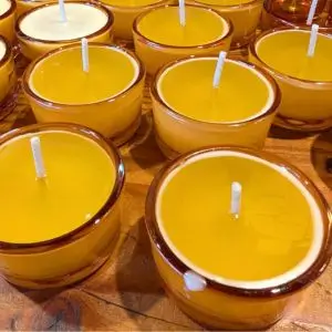 A close-up of several small amber glass candles filled with bright golden yellow wax. Each candle has a centered white wick, and they are arranged neatly on a wooden surface. One candle in the foreground has a small drip of wax on its rim.