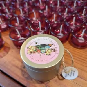 A gold tin candle with a pink label featuring a songbird, bee, and floral illustration, branded “The Songbird and The Bee – Blue Mountains.” The tin is placed on a wooden surface in front of rows of red glass votive candles with white wicks. A round tag attached to the tin reads “Lemon Verbena.”