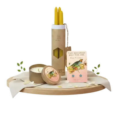 A wooden tray display featuring yellow taper candles in a tube, a round tin candle, and blue flower stud earrings on a card, decorated with white petals.
