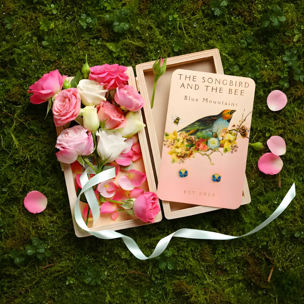 blue flower earrings on a card on a box filled with roses on placed on top of mossy ground