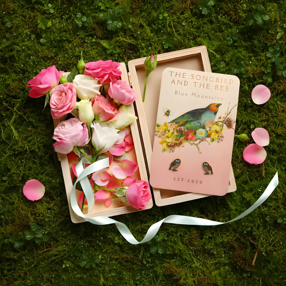 owl earrings on a card on a box filled with roses on placed on top of mossy ground